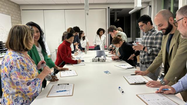 A group of adults take part in a workshop. Standing around a table, they discuss an exercise.