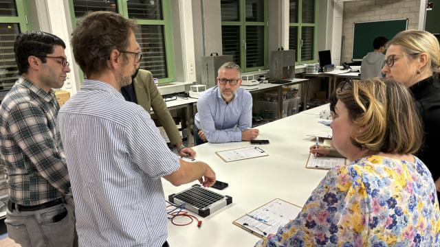 A group of adults take part in a workshop. Standing around a table, they discuss an exercise.