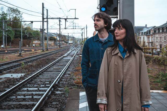 Ein Mann und eine Frau stehen auf dem Bahnsteig eines Bahnhofs und blicken nach links.