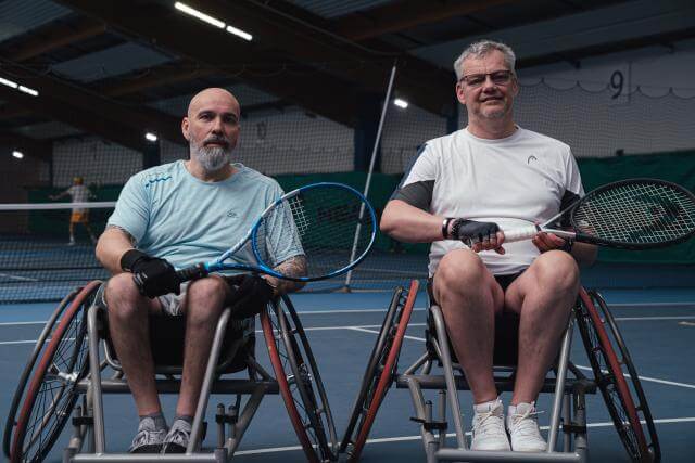 Two men in wheelchairs in a tennis hall, each holding a tennis racket.