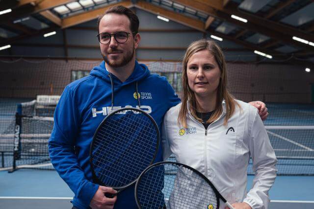 A man and a woman pose for a photo in a tennis hall. The man on the left is holding a racket.