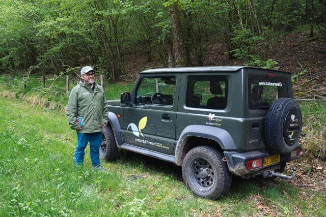 A man standing to the left of a Natur & Ëmwelt vehicle, parked on a lawn in the middle of nature.
