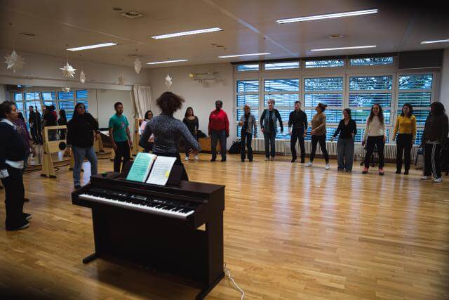 Vue d’une grande salle de répétition avec un piano au premier plan et un groupe de chanteurs en demi-cercle à l’arrière-plan.