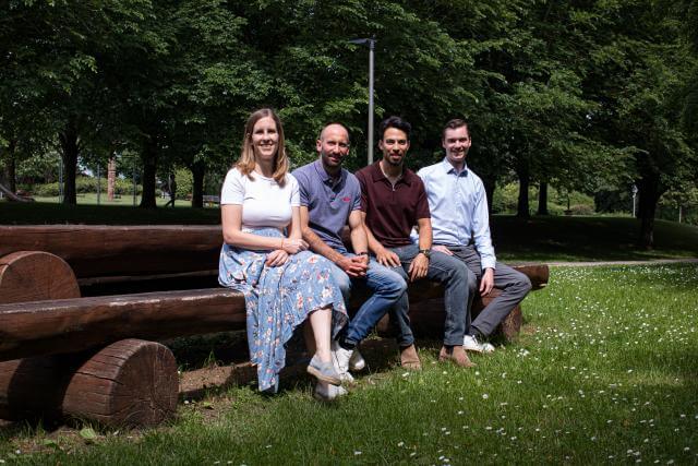 eine Frau udn drei Männer sitzen auf einer Bank auf einer Wiese