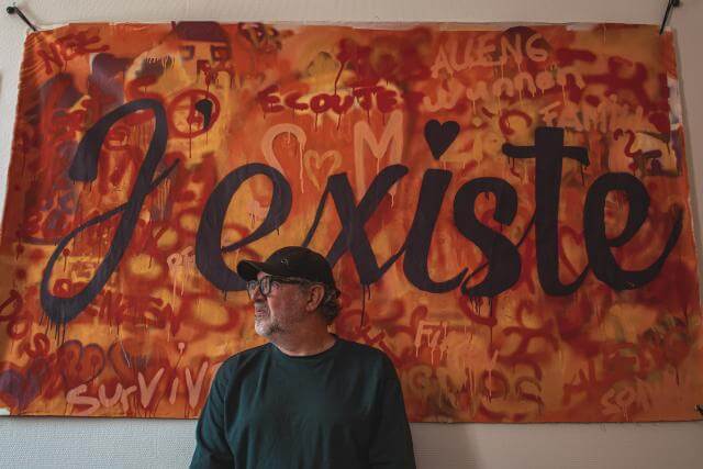 Porträt eines Mannes vor einem Plakat mit der Aufschrift „Ich existiere“.