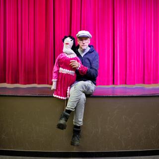 portrait de Pit Vinandy, assis sur une scène devant un rideau rouge. Il tient une marionnette dans la main