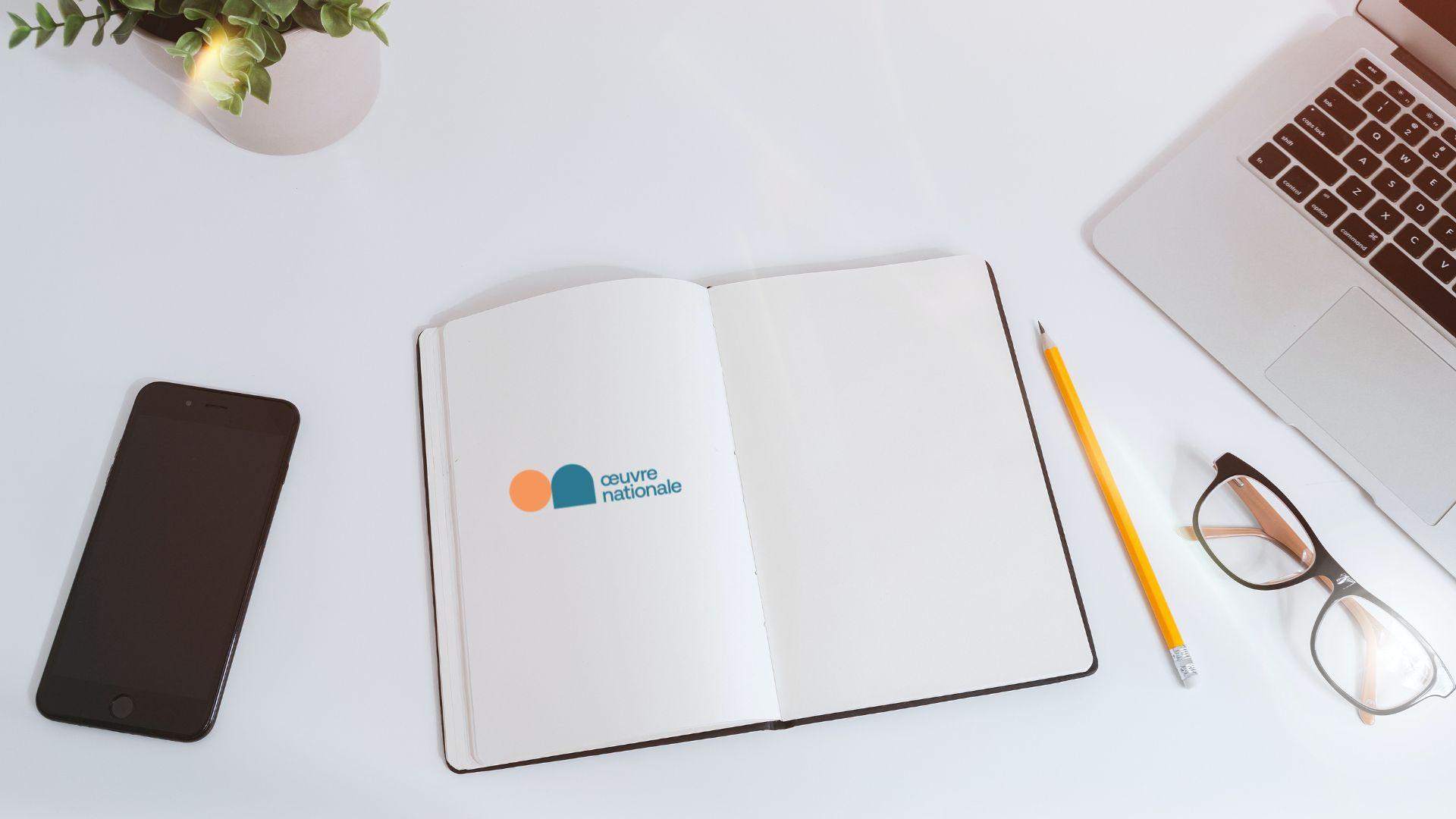 Foto eines Tisches, auf dem ein offenes Notizbuch mit dem Logo der Œuvre, ein Smartphone und ein Laptop zu sehen sind