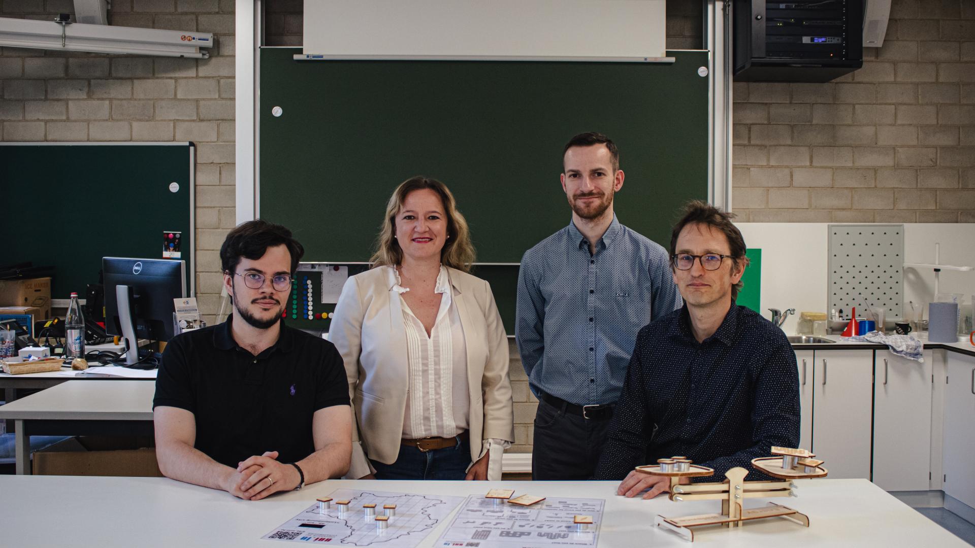A group of 3 men and a woman in a classroom that looks like a science laboratory. They stand in front of a blackboard and look straight into the camera.