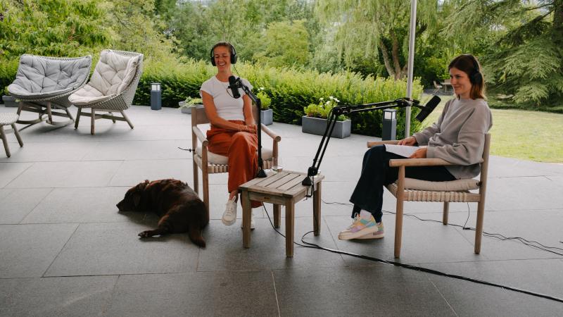 tournage d'un podcast, deux femmes sont assises sur une terrasse avec un chien, elles portent des casques et des micros se trouvent devant elles.