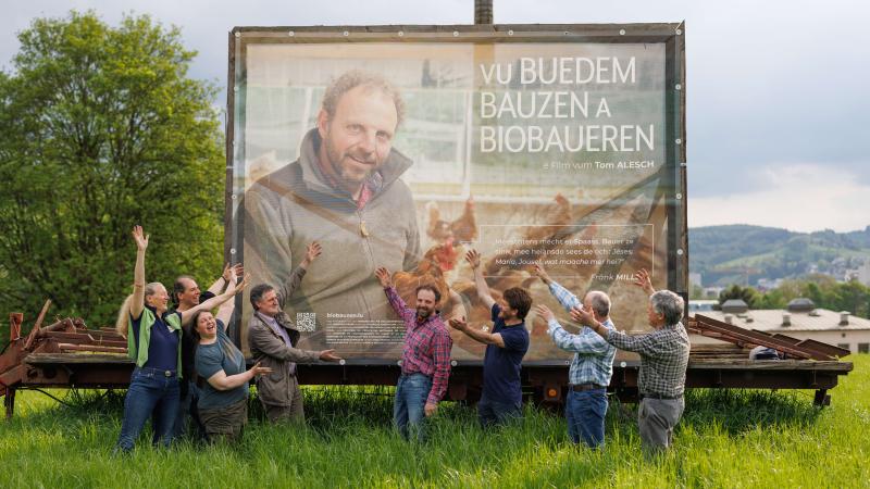 Personnes dans un pré devant un grand panneau sur lequel est montré une image du film "vu Buedem, Bauzen a Biobaueren"