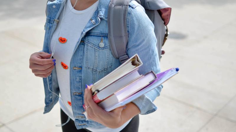 jeune étudiante vétue d'un blouson en jean et tenant des livres scolaires à la main. Elle porte un sac à dos et des écouteurs avec fils.