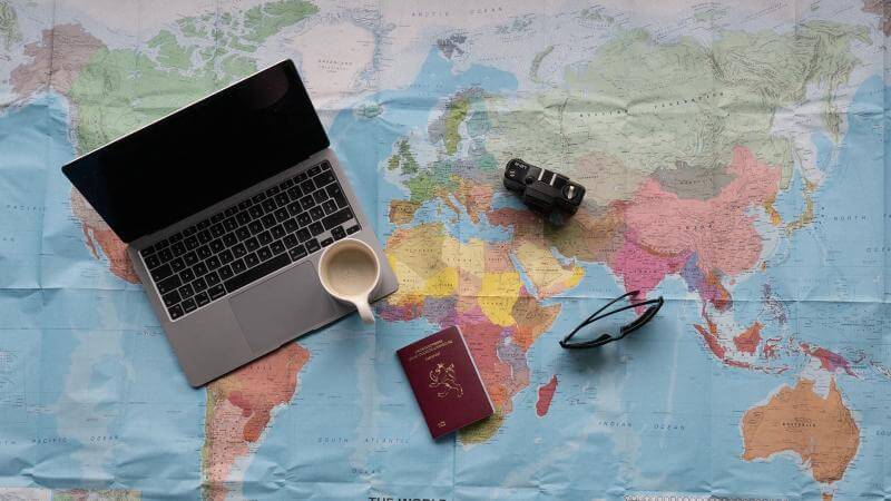 Une photo d'une carte du monde. Dessus sont posés un ordinateur, un passeport, une caméra et des lunettes de soleil.