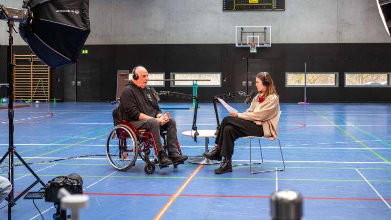 Set-up des Podcasts in einer Sporthalle. Links sitzt ein Mann in einem Rollstuhl vor seinem Mikrofon, vor ihm eine Frau, die ebenfalls vor einem Mikrofon sitzt.