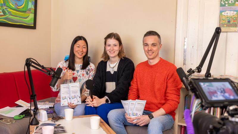 Zwei Frauen und ein Mann sitzen auf einem Sofa, man sieht eine Kamera und Mikrofone, sind sind dabei einen Podcast auszuzeichnen