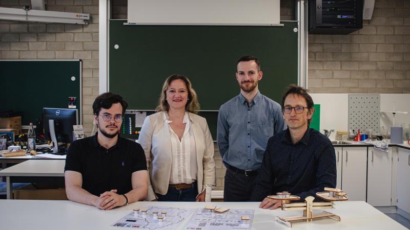 A group of 3 men and a woman in a classroom that looks like a science laboratory. They stand in front of a blackboard and look straight into the camera.