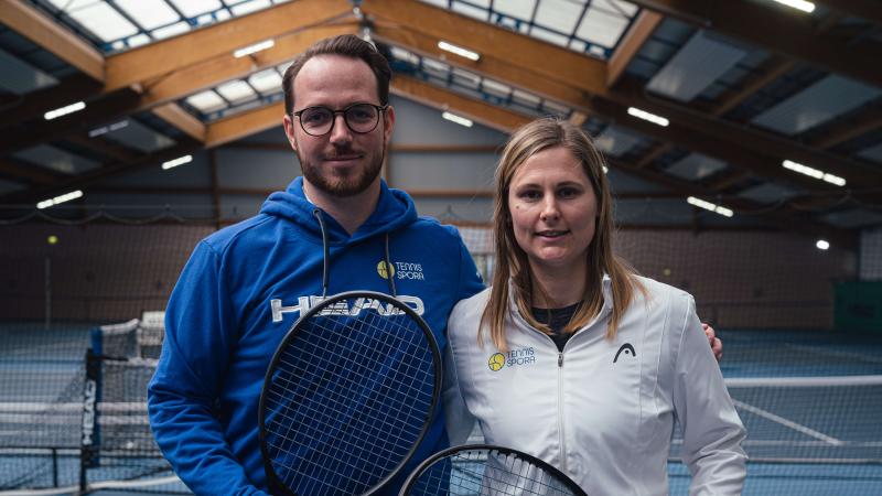 Un homme et une femme posent pour la photo dans un hall de tennis. L’homme, situé à gauche, tient une raquette.