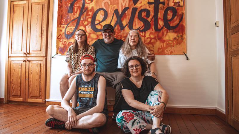 Photo of a group of people sitting in front of a poster with “I exist” written on it.